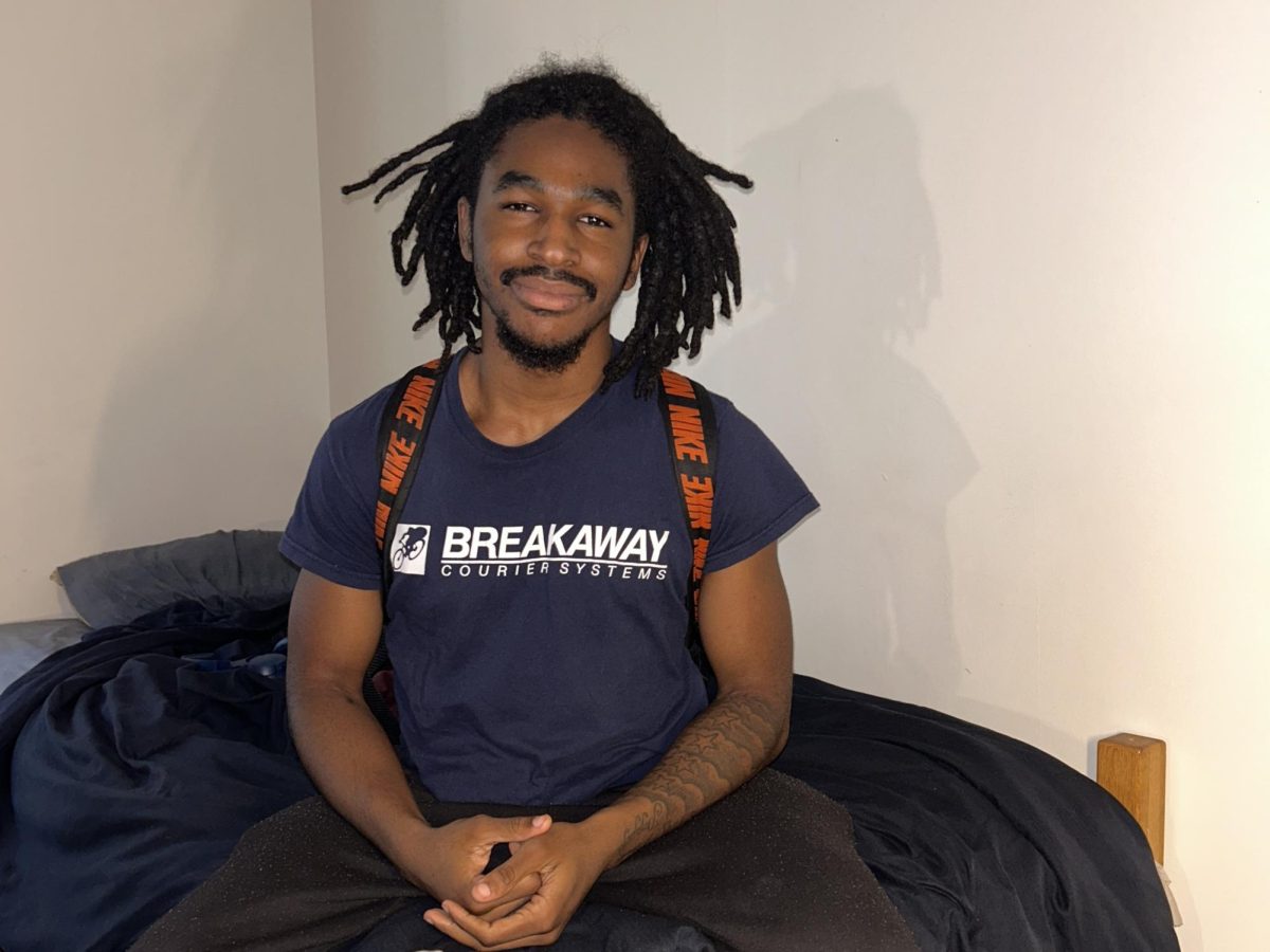 Hamadi Martin, a junior at SUNY Old Westbury, sits on his dorm bed in Woodlands Hall on March 25, 2026, wearing a shirt from his father’s company, Breakaway Courier Systems. PHOTO BY: JULIA WALTER